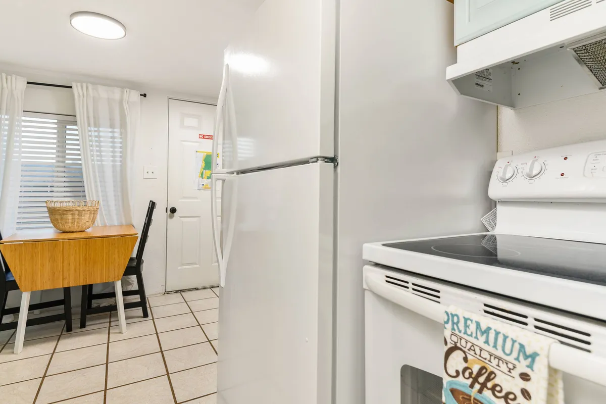 A warm, lived-in kitchen scene showing a table with a bowl, a white refrigerator, and a stove with a coffee towel. The setting suggests a place where guests—and their pets—can gather, prepare meals, and enjoy time together.