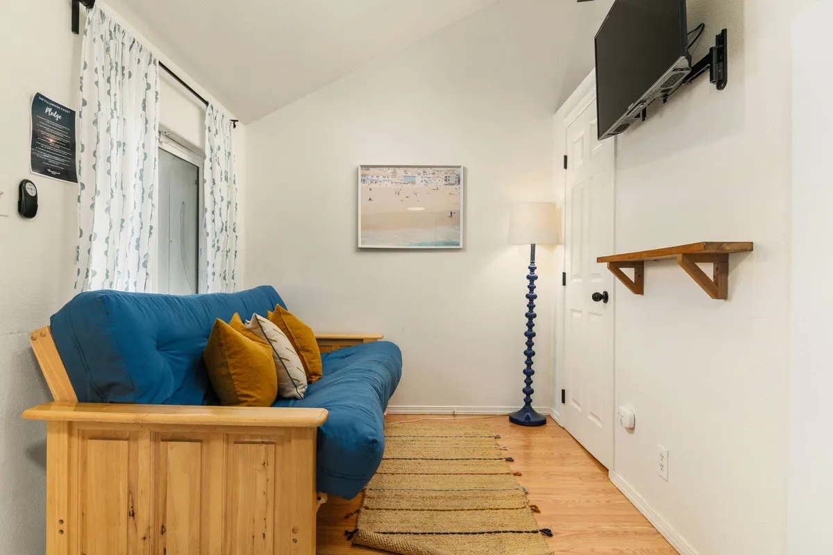 A cozy living area with a blue futon, colorful throw pillows, a blue lamp, wall art, and a mounted TV. The arrangement is perfect for movie nights, reading, or socializing, and the décor adds a playful, coastal touch.