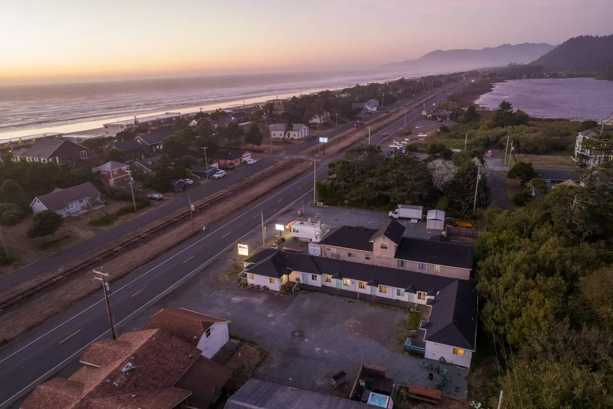 The image captures a tranquil view of the Oregon coast from the perspective of the Sea Haven Motel. The Pacific Ocean glimmers in the background, with a quiet road leading towards the waves, flanked by charming coastal buildings and majestic forested mountains. The scene invites visions of relaxed beach walks, sunset watching, and easy access to both nature and the heart of Rockaway Beach.