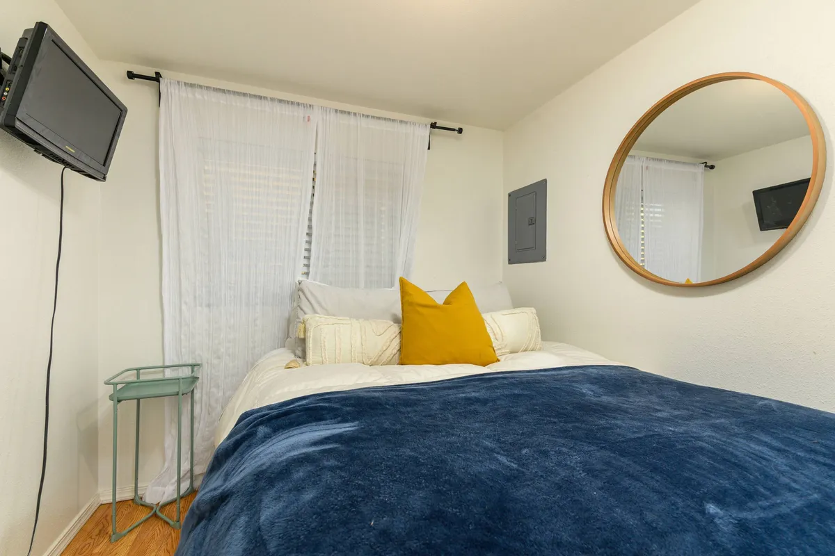 A cozy bedroom scene featuring a plush queen bed adorned with a blue blanket and a vibrant yellow accent pillow. Soft curtains filter the natural light, and a round mirror above the bed adds a touch of elegance. There is a TV placed for convenient viewing from the bed, contributing to a relaxing and welcoming atmosphere.