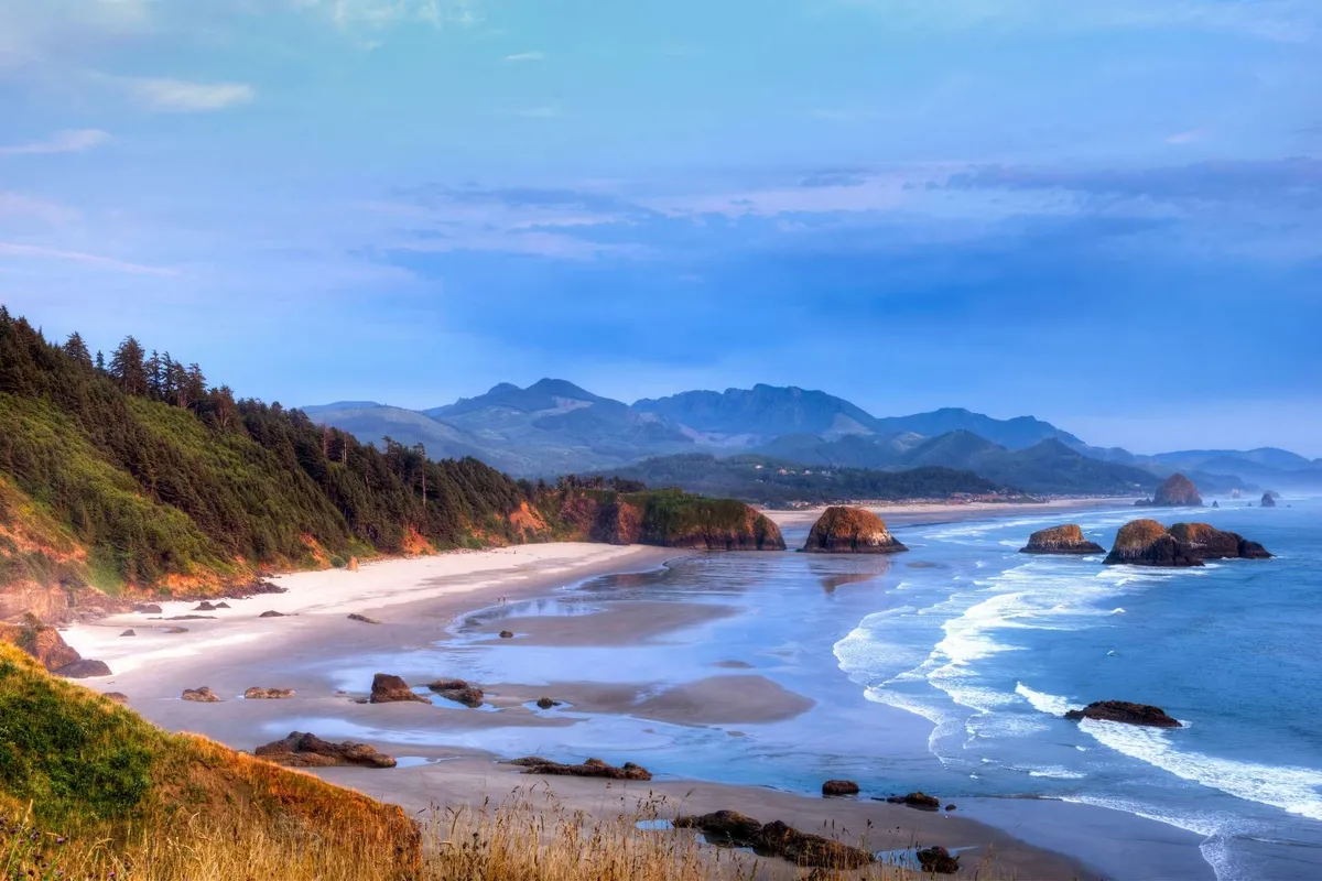 An expansive view of the sandy Rockaway Beach coastline with gentle waves lapping ashore. Distant mountains rise on the horizon, enhancing the sense of natural beauty and openness. The foreground shows the sandy beach, perfect for strolling, picnicking, or letting kids and pets play.