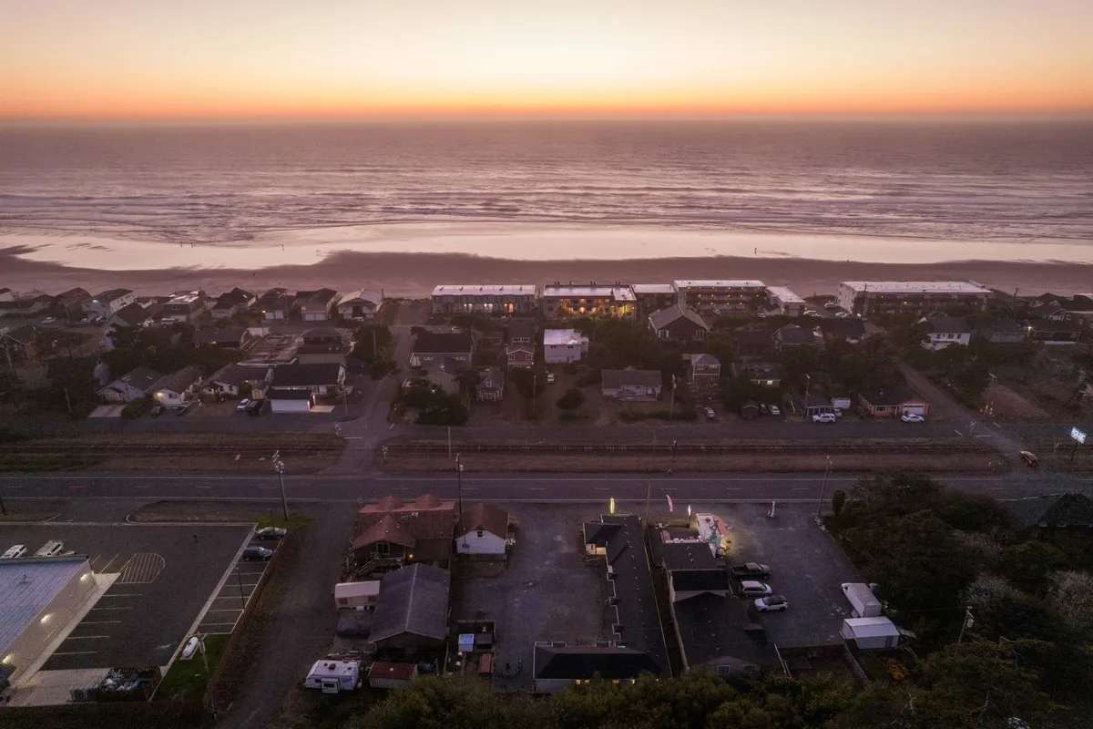 This image captures a peaceful evening scene overlooking Rockaway Beach. The ocean waves shimmer under a sunset sky, with rows of houses lining a quiet road that leads directly toward the beach. The scene feels inviting, with a sense of community and the promise of easy access to the coast.